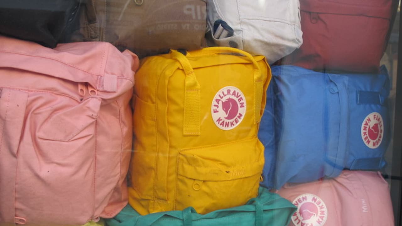 Colorful bags stacked together, various fabric backpacks in pink, yellow, blue, maroon, and white, visible brand logo on yellow and blue bags, arranged closely in a display setting, likely for sale or storage