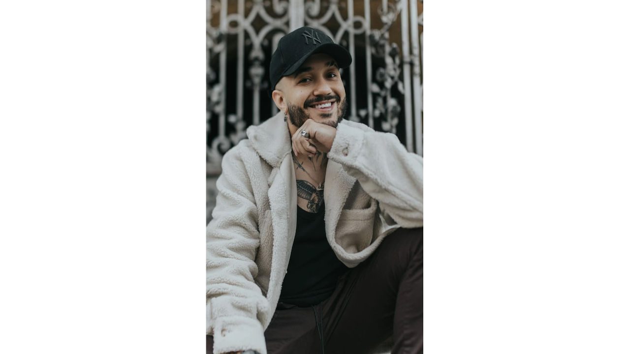 Smiling man in a beige fleece jacket and black cap sitting in front of an ornate gate