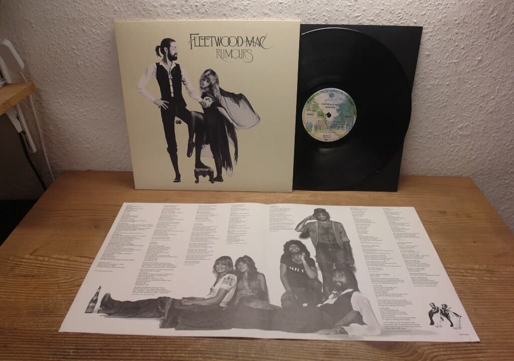 Fleetwood Mac – Rumours album cover, beige background, Mick Fleetwood in black outfit holding a cane, Stevie Nicks in flowing dress posing with one foot on a stool, vintage 1977 vinyl record artwork