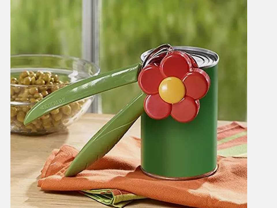 Decorative flower-shaped can opener, red petals with yellow center, green handles resembling leaves, attached to a green tin can, placed on a wooden table with a glass bowl of beans in the background, near a window