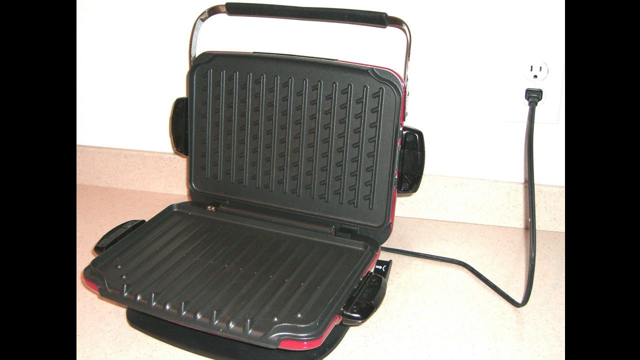Open George Foreman electric grill with ridged non-stick cooking plates, plugged into wall outlet, placed on a kitchen countertop