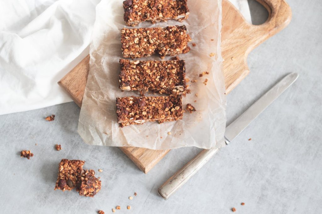 Four granola bars on parchment paper, placed on wooden board, one broken piece on gray surface, crumbs scattered