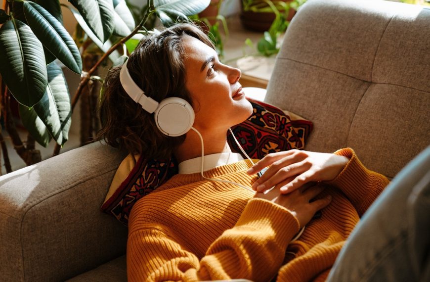 Woman laying on couch with headphones on smiling and staring out the window in the sunlight