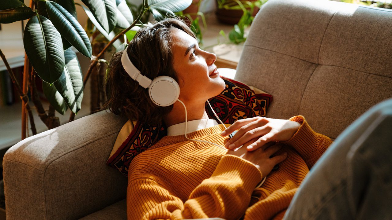 Woman laying on couch with headphones on smiling and staring out the window in the sunlight