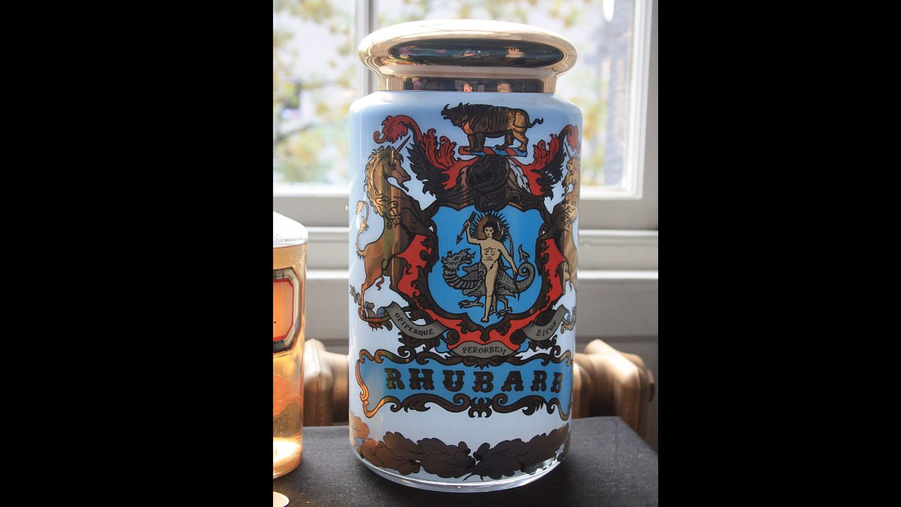 historic apothecary jar labeled “Rhubarb,” with ornate crest design featuring a mythological figure, lion, unicorn, dragon, and gold lid, displayed near a window