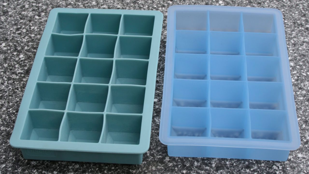 Two silicone ice cube trays, one teal and one light blue, each with 15 square compartments, placed on a speckled countertop