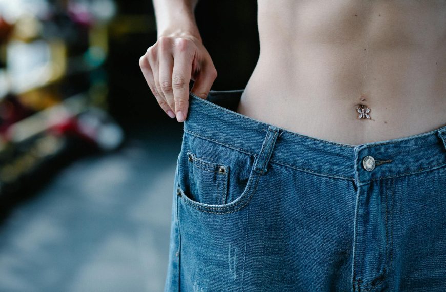 Person wearing oversized blue jeans, holding waistband to show looseness, bare midriff visible, background slightly blurred with indoor shelving, suggests weight loss or ill-fitting clothes