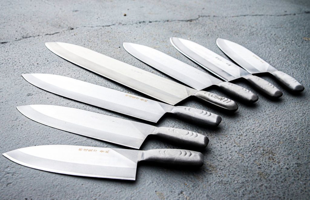 Set of seven stainless steel kitchen knives, arranged in descending size order, on textured gray surface