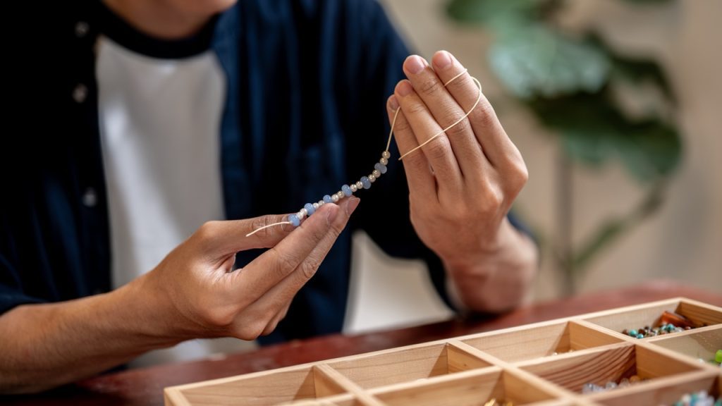 Young man artist holding and looking at ceramic beads on thread while making a bracelet in a class or studio.
