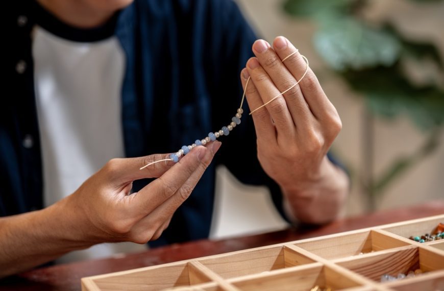 Young man artist holding and looking at ceramic beads on thread while making a bracelet in a class or studio.