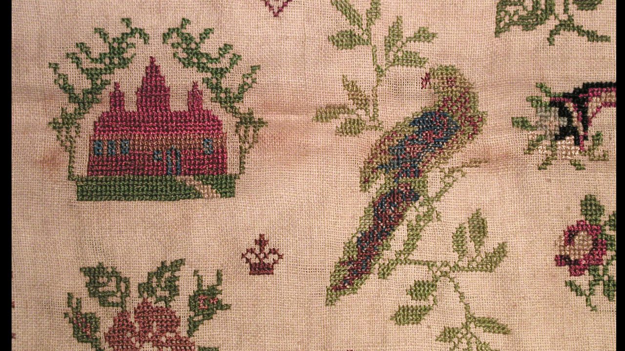 Vintage needlepoint sampler on beige fabric featuring a red house, green trees, a colorful bird, and floral motifs