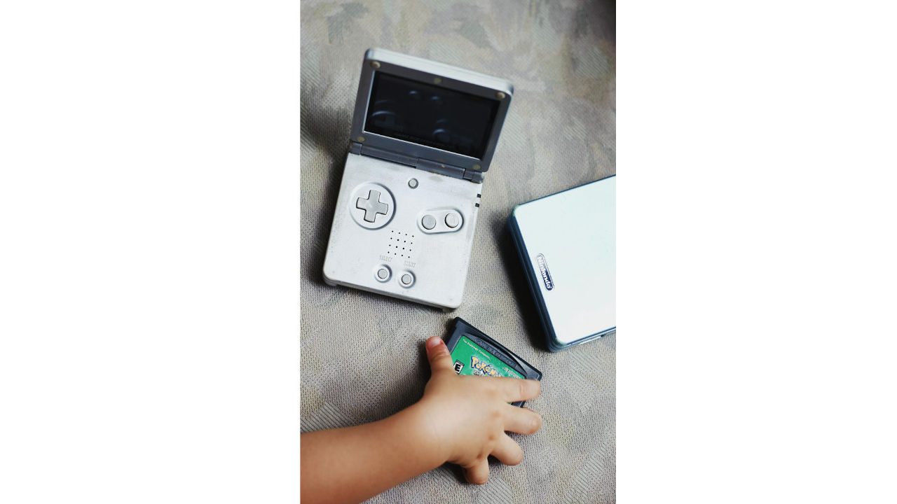 Child’s hand holding a Pokémon game cartridge near a silver Nintendo Game Boy Advance SP on a fabric surface