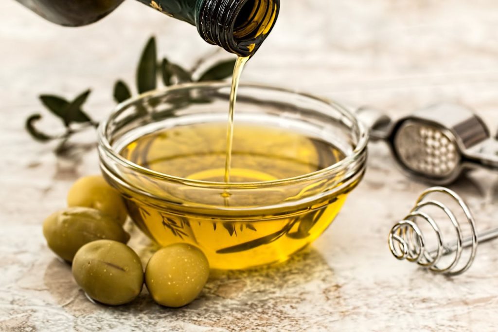 Olive oil being poured into a glass bowl with green olives beside it, whisk and garlic press nearby