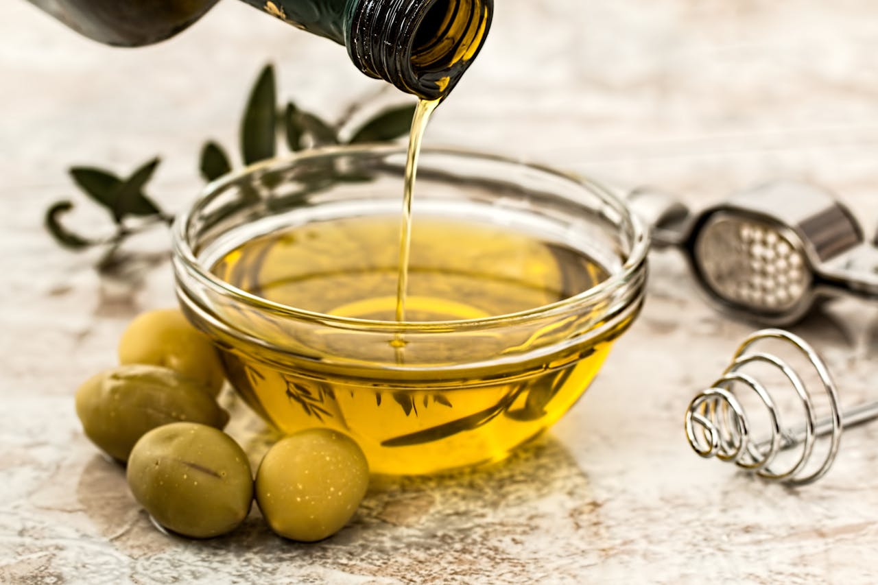 Olive oil being poured into a glass bowl with green olives beside it, whisk and garlic press nearby