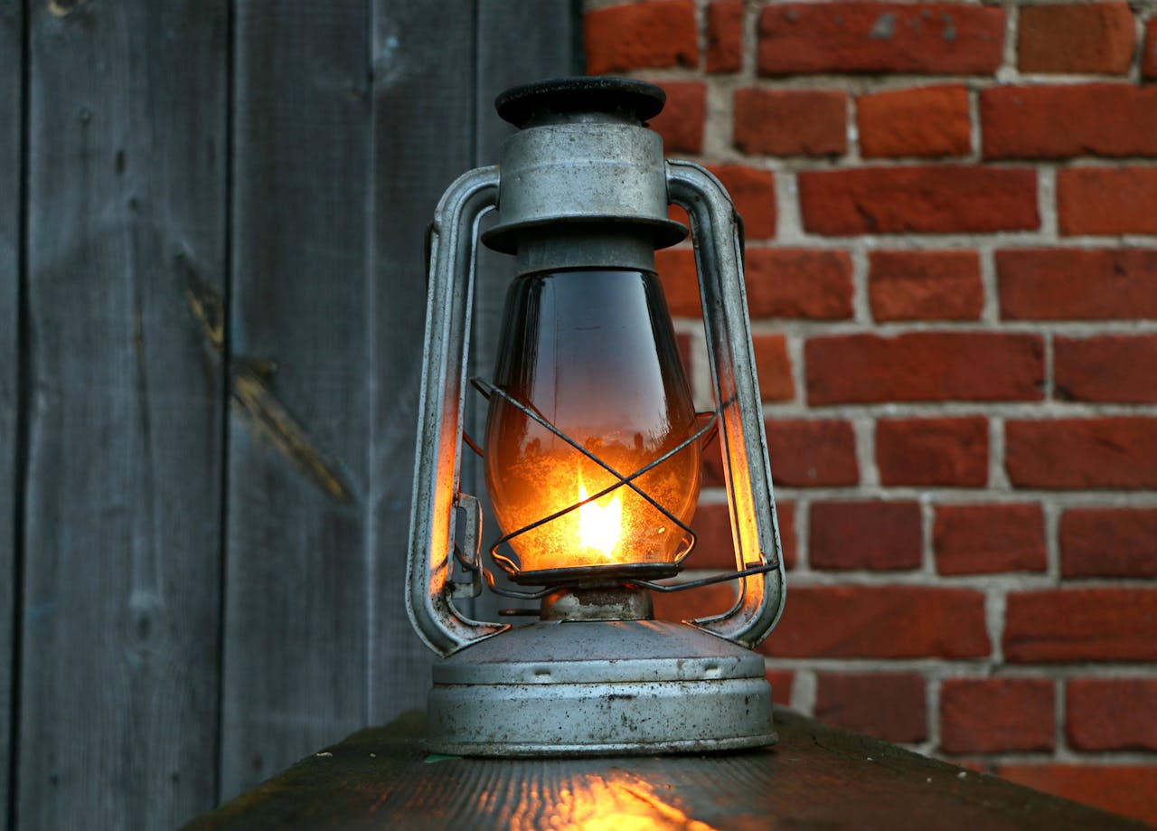 Lit vintage oil lamp, metal frame, glowing glass, placed on wooden surface, brick and wood wall background