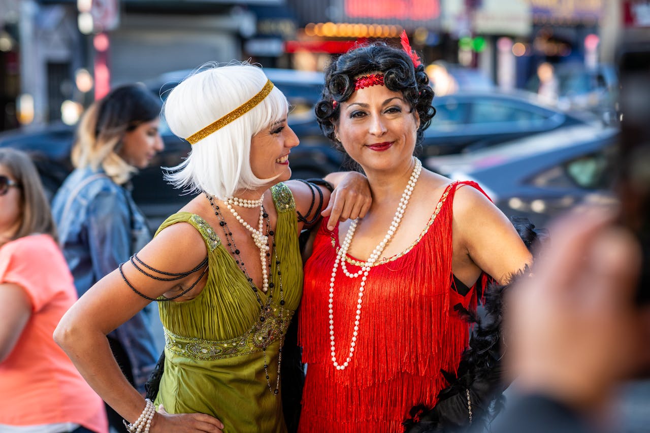 Two women in 1920s flapper costumes smiling outdoors, wearing fringe dresses, pearls, and headbands