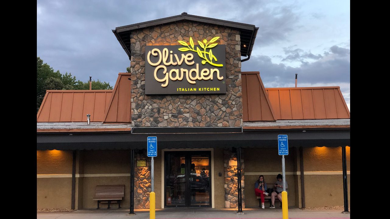 Olive Garden restaurant with stone facade, orange roof, and glowing sign at dusk with people sitting outside