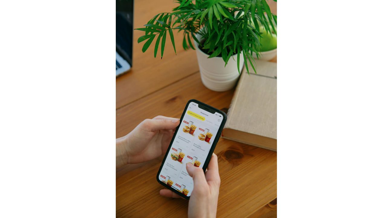Person browsing fast food meal options on smartphone, online food delivery app open, wooden desk background