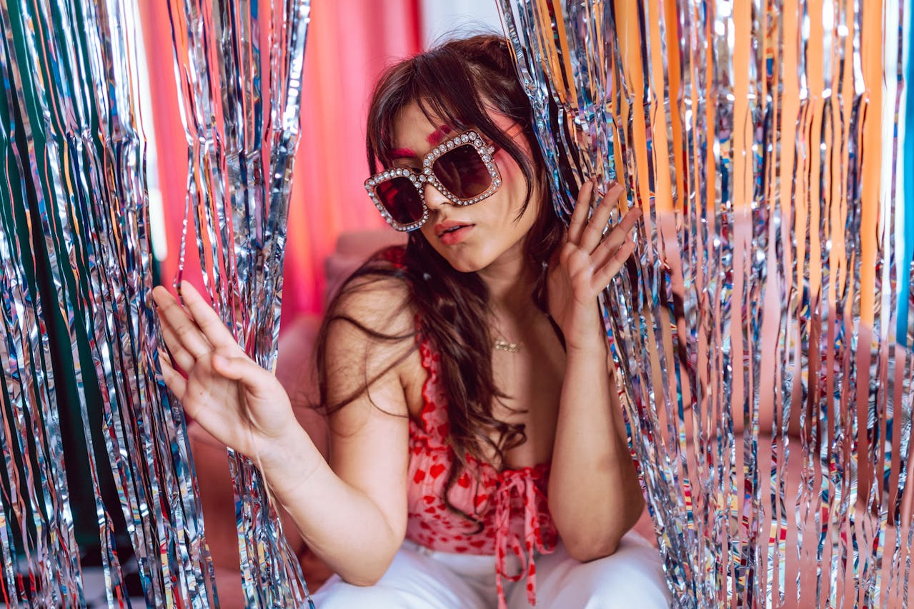 Woman wearing oversized rhinestone sunglasses and red outfit posing behind silver and orange party streamers