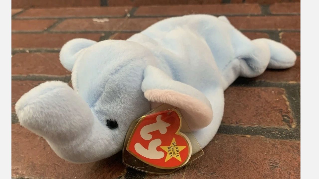Light blue TY Beanie Baby elephant, floppy ears, trunk curled, heart tag attached, lying on brick surface