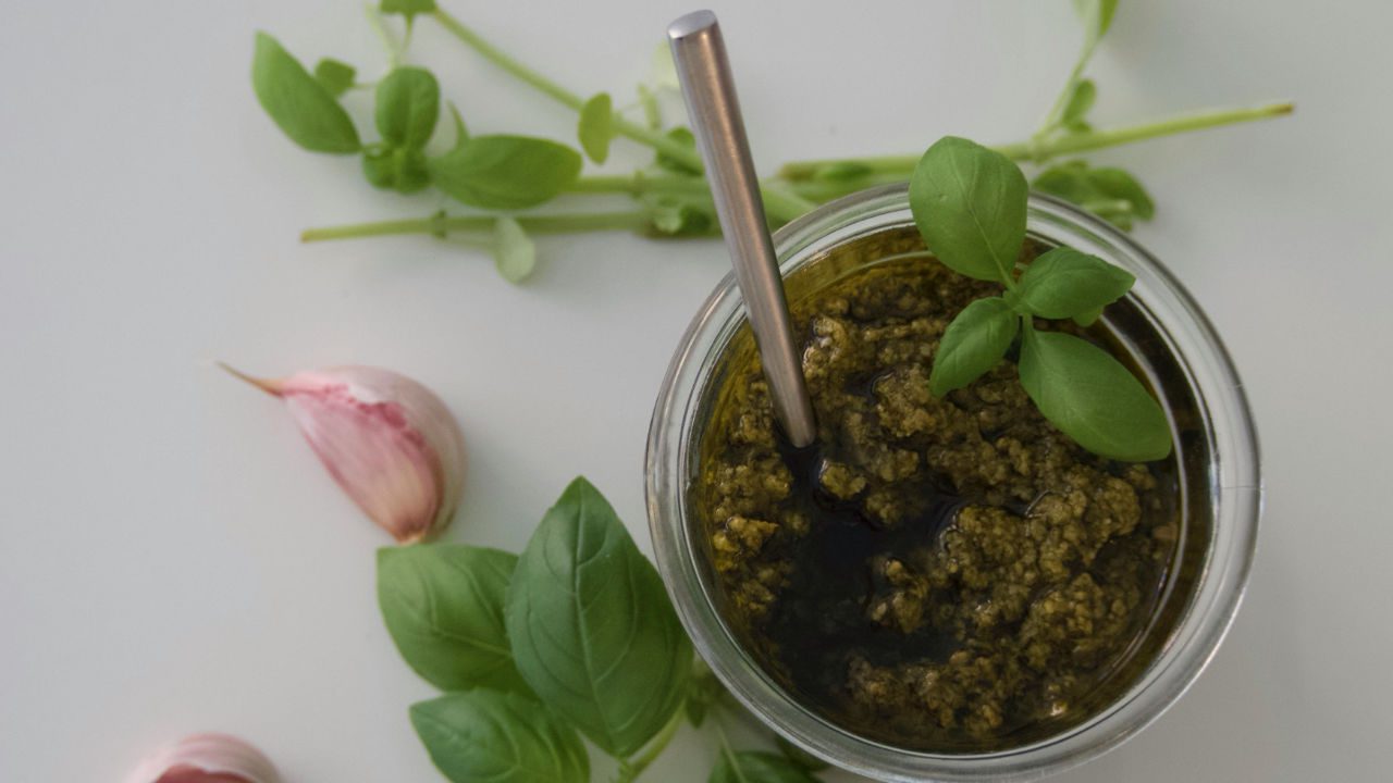 Glass jar of green pesto sauce with spoon, basil leaves on top, garlic cloves and fresh herbs on white background