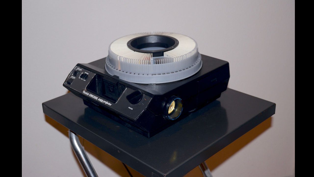 Vintage photo slide carousel projector with circular slide tray, placed on a small metal stand