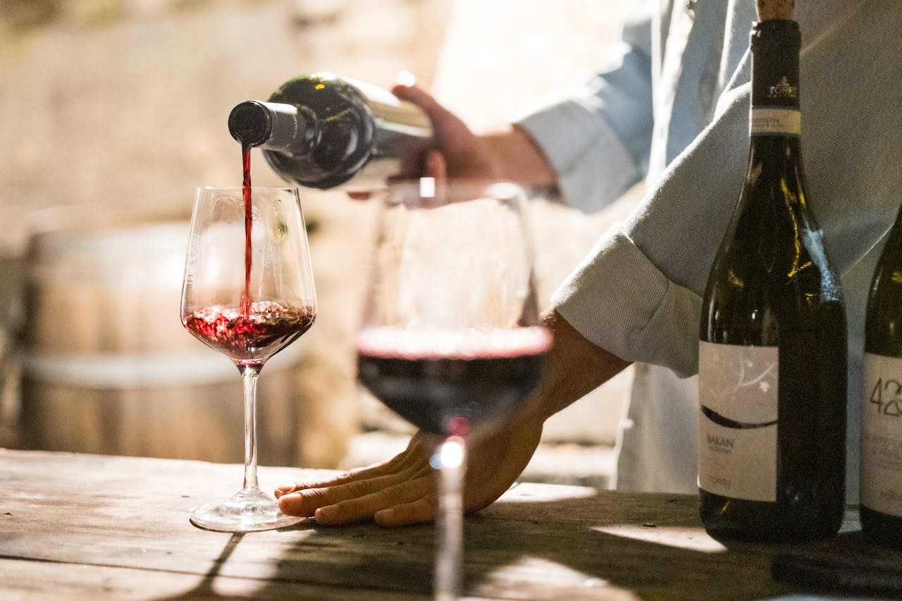 Person pouring red wine into a wine glass beside other filled glasses and bottles on a rustic table