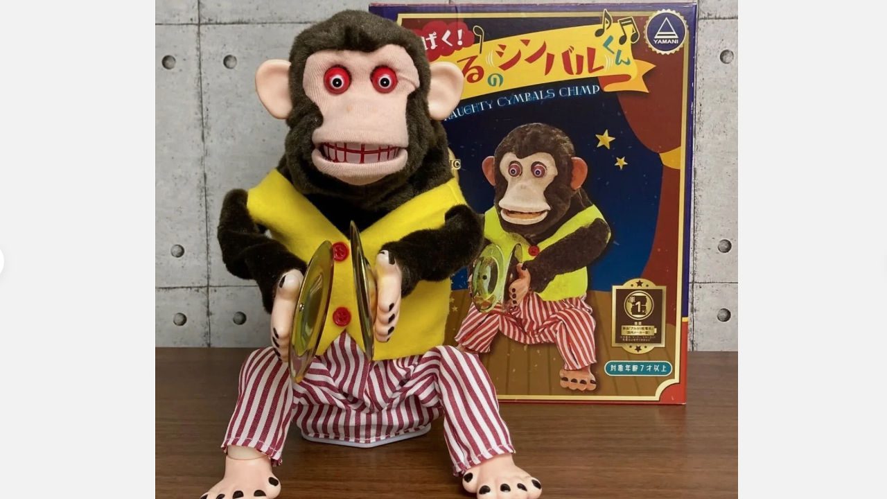 vintage cymbal-banging monkey toy with yellow vest, red-striped pants, red eyes, seated on table, original box in background