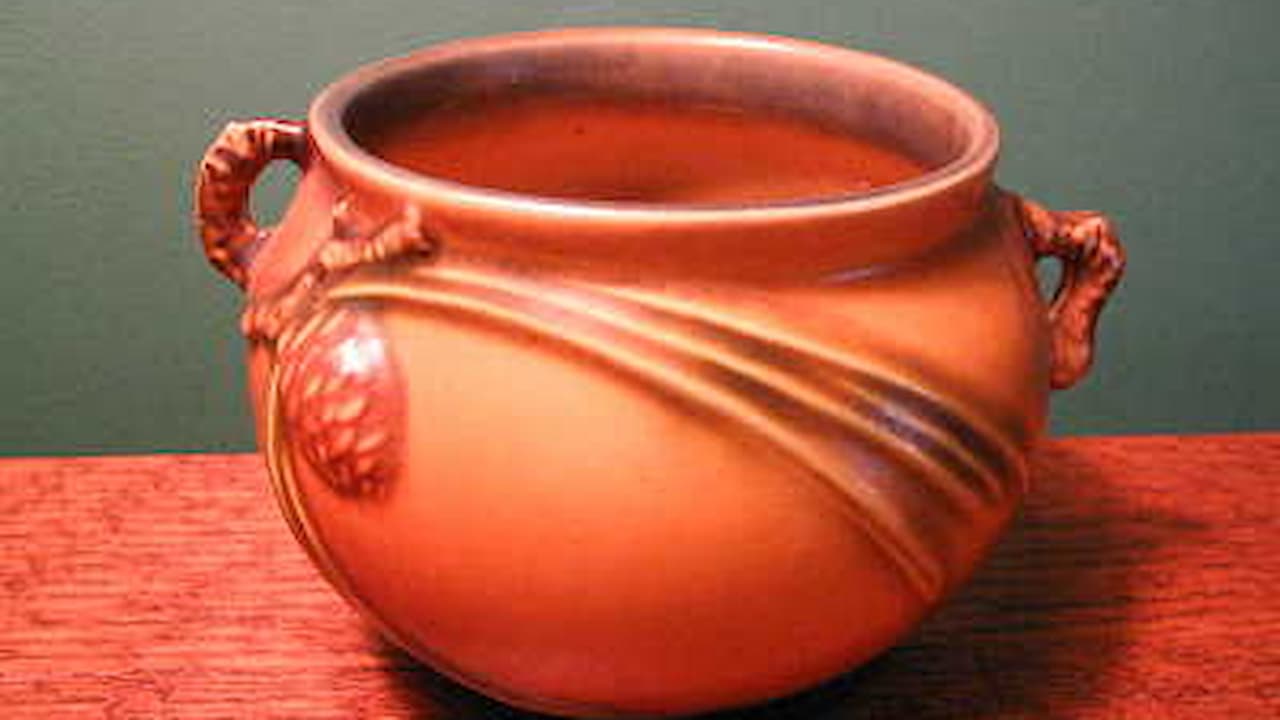 Orange ceramic pot with two side handles, decorative rope-like design wrapped around the body, matte finish, placed on a red textured surface, green background