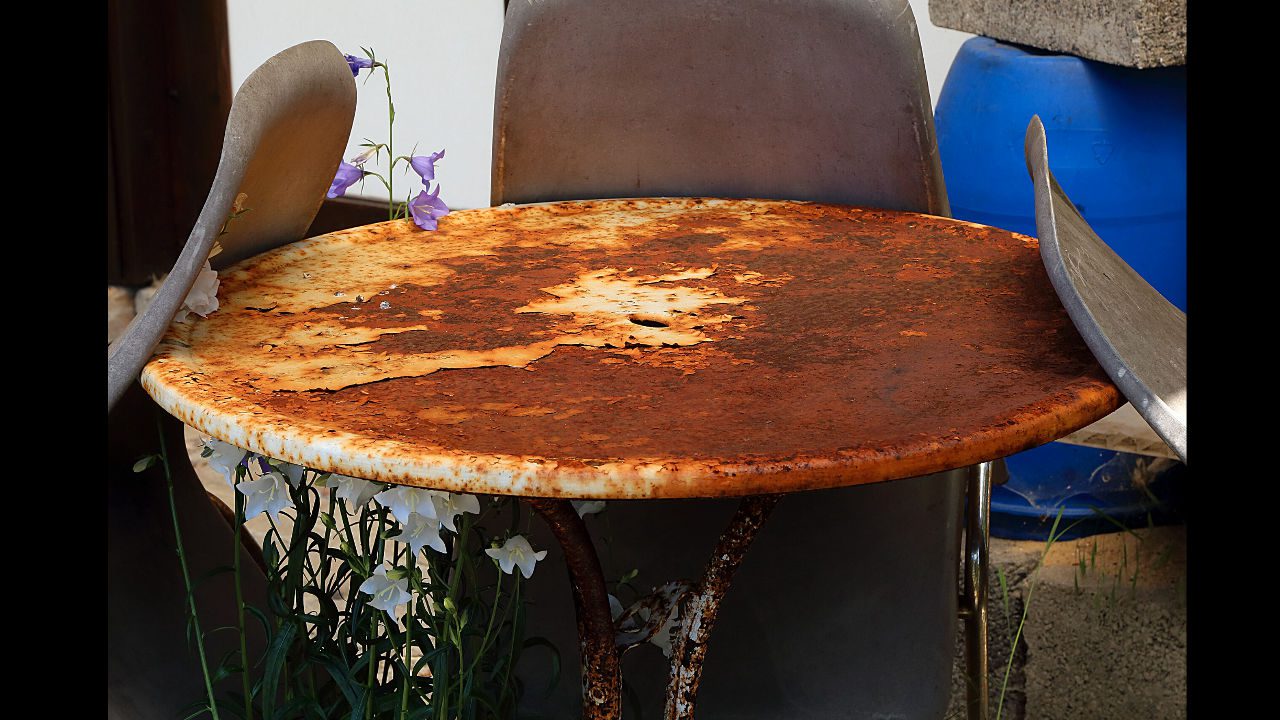 Rusty round metal table surrounded by worn chairs and wildflowers