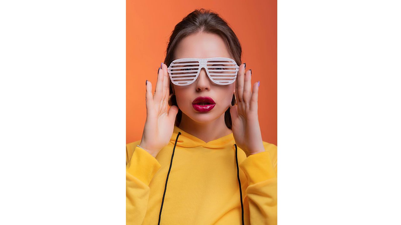 Woman in yellow hoodie wearing white shutter shades, bright lipstick, posing with hands near face, orange background