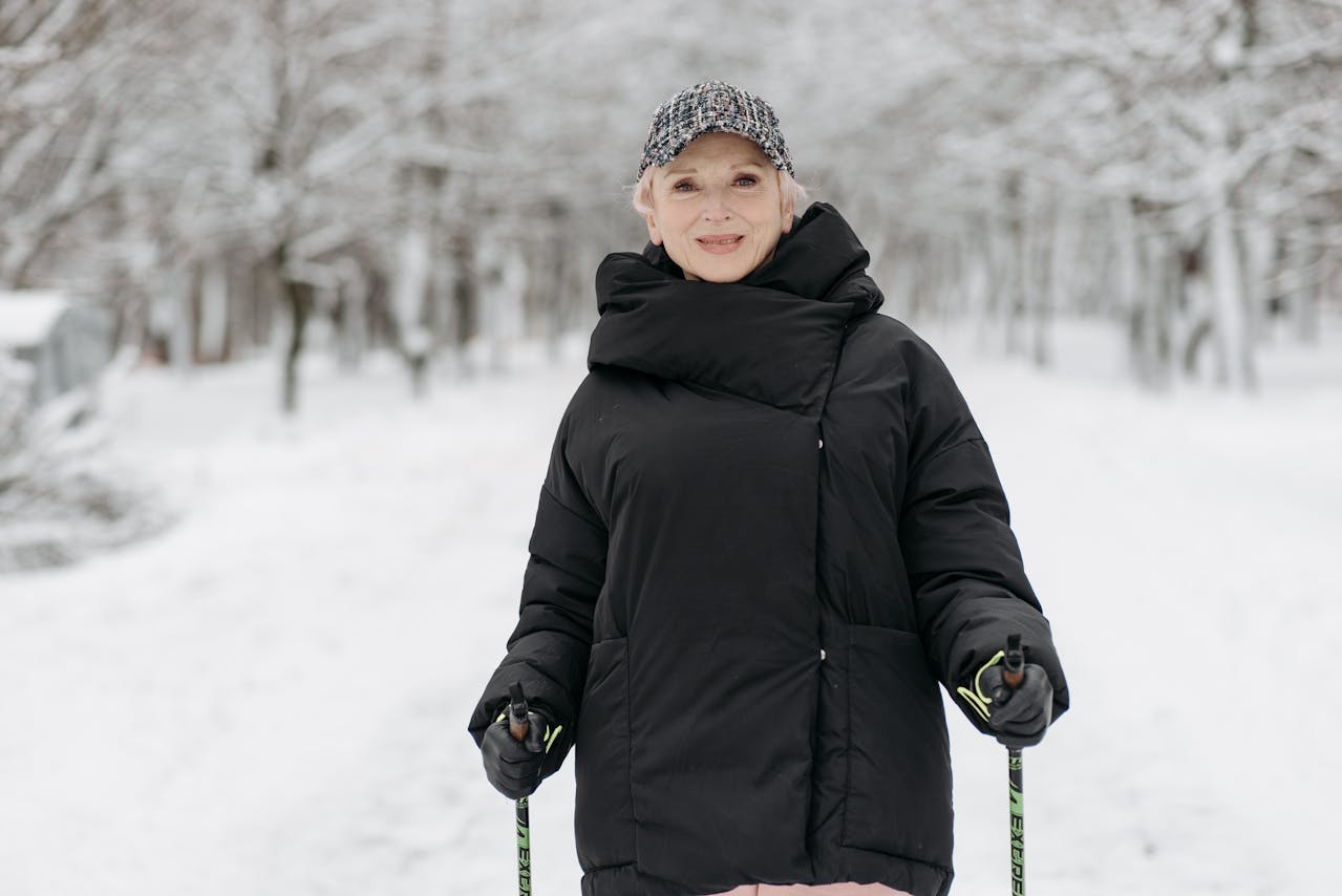 Older woman wearing black ski jacket and winter cap walking with poles in snowy park