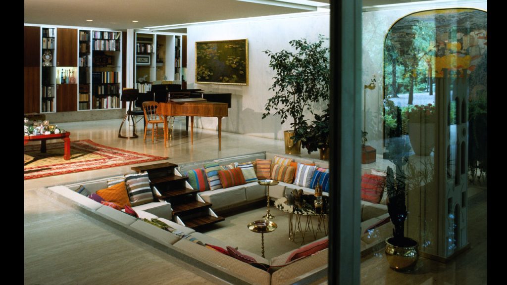 Retro sunken living room with colorful pillows, built-in seating, and mid-century modern decor, including a piano and bookshelf