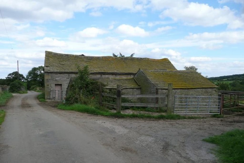 old barn, sagging roof, moss-covered tiles, weathered stone walls, rural dirt road, wooden fence, overgrown vegetation, cloudy sky