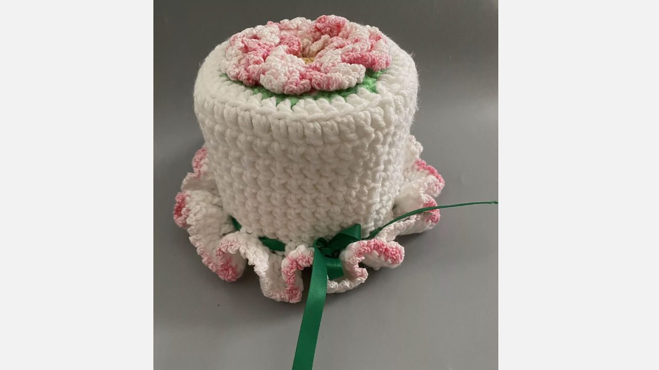 Crocheted toilet paper cover with a white body, pink flower top, ruffled trim, and green ribbon