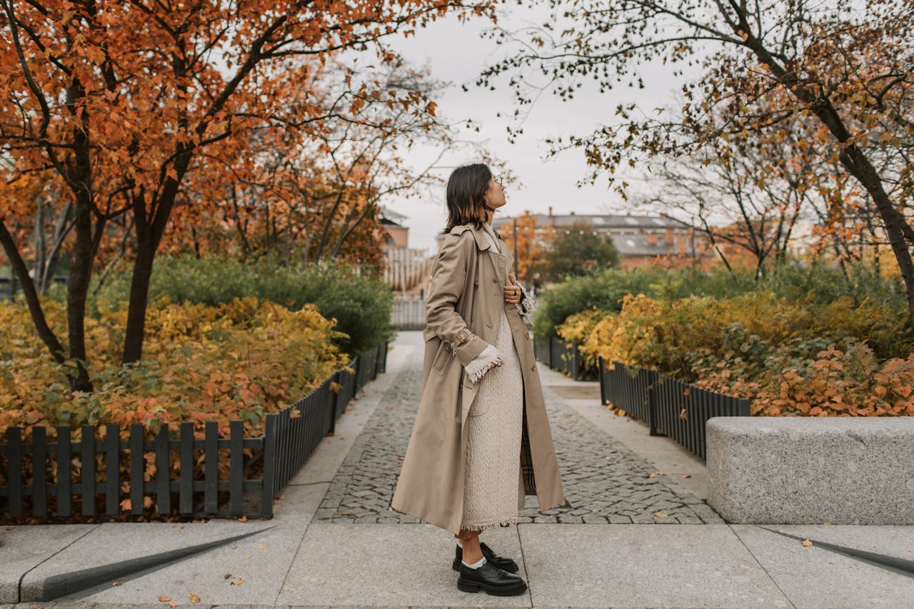 Woman wearing a beige trench coat and dress standing in a park with autumn-colored trees