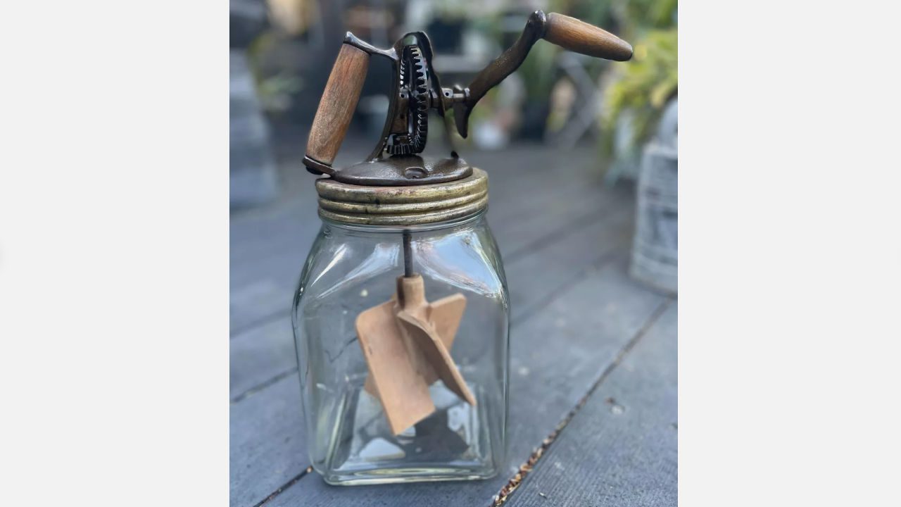 Vintage glass butter churn with hand crank, wooden paddles, and metal gear top sitting on wooden deck