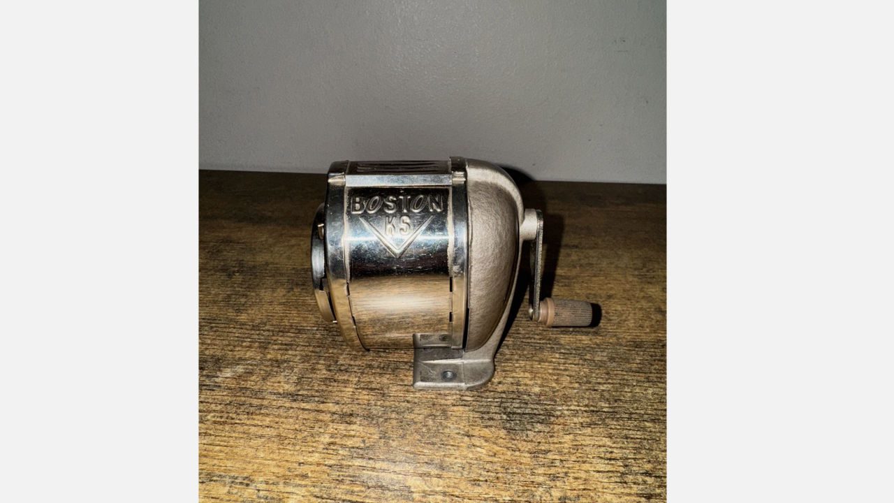 Vintage wall-mounted metal pencil sharpener labeled "BOSTON KS" with hand crank and mount base