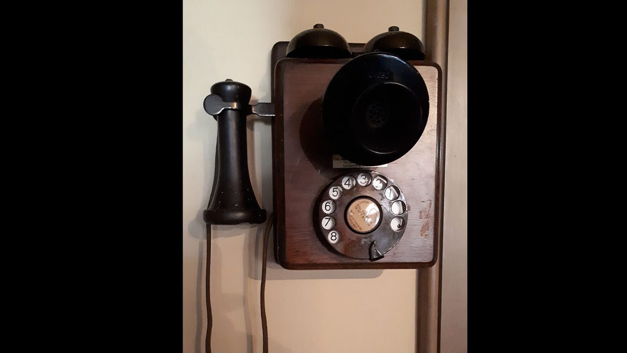 Vintage wall-mounted rotary dial telephone, wooden body, black earpiece, twin metal bells on top, mounted on beige wall