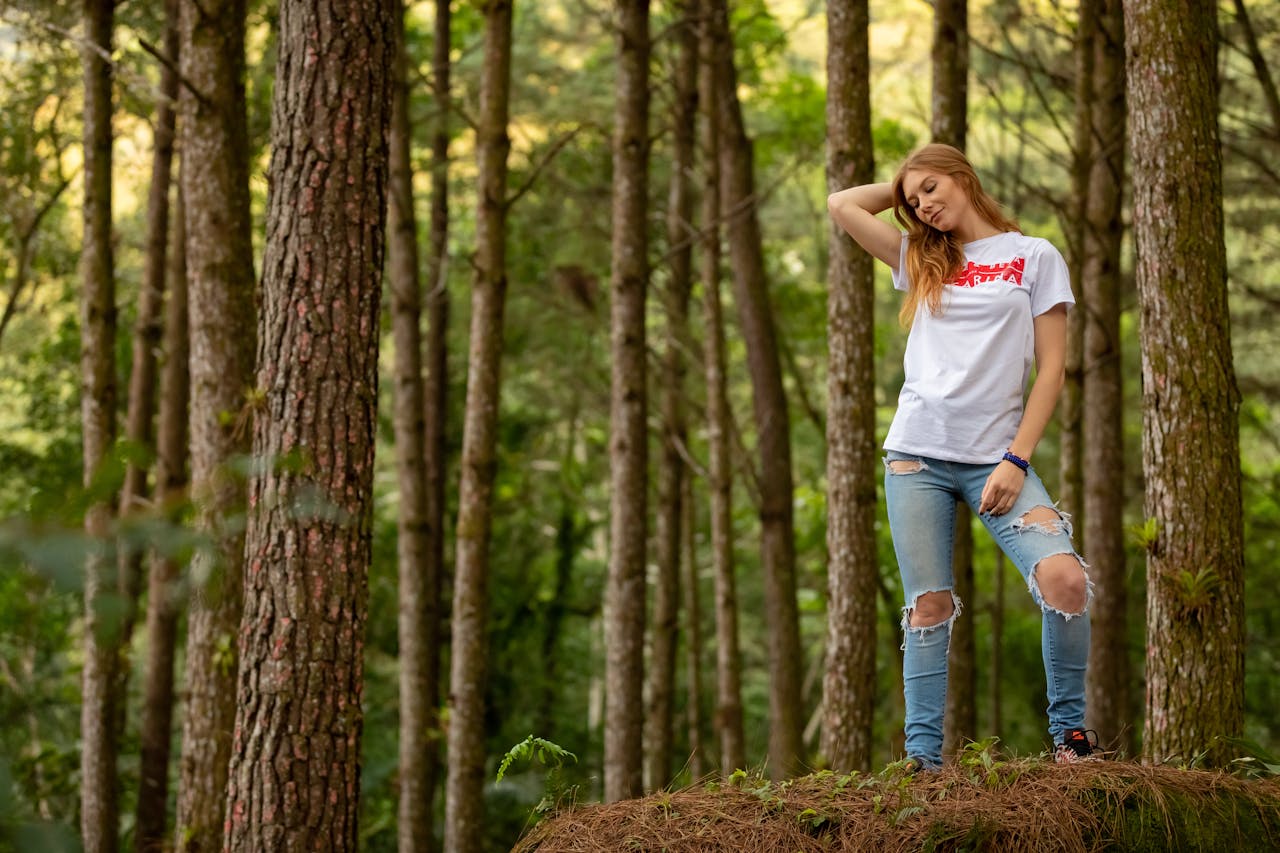 Woman wearing white t-shirt and ripped skinny jeans standing in a forest, hand resting on head, natural light