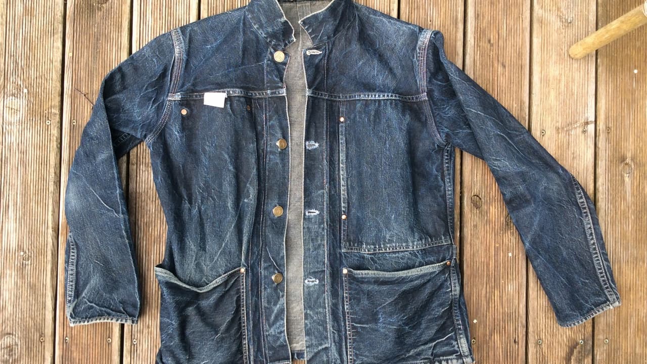 Worn denim work jacket, button-up front, multiple front pockets, faded and distressed areas, laid flat on wooden surface, long sleeves, visible stitching and seams