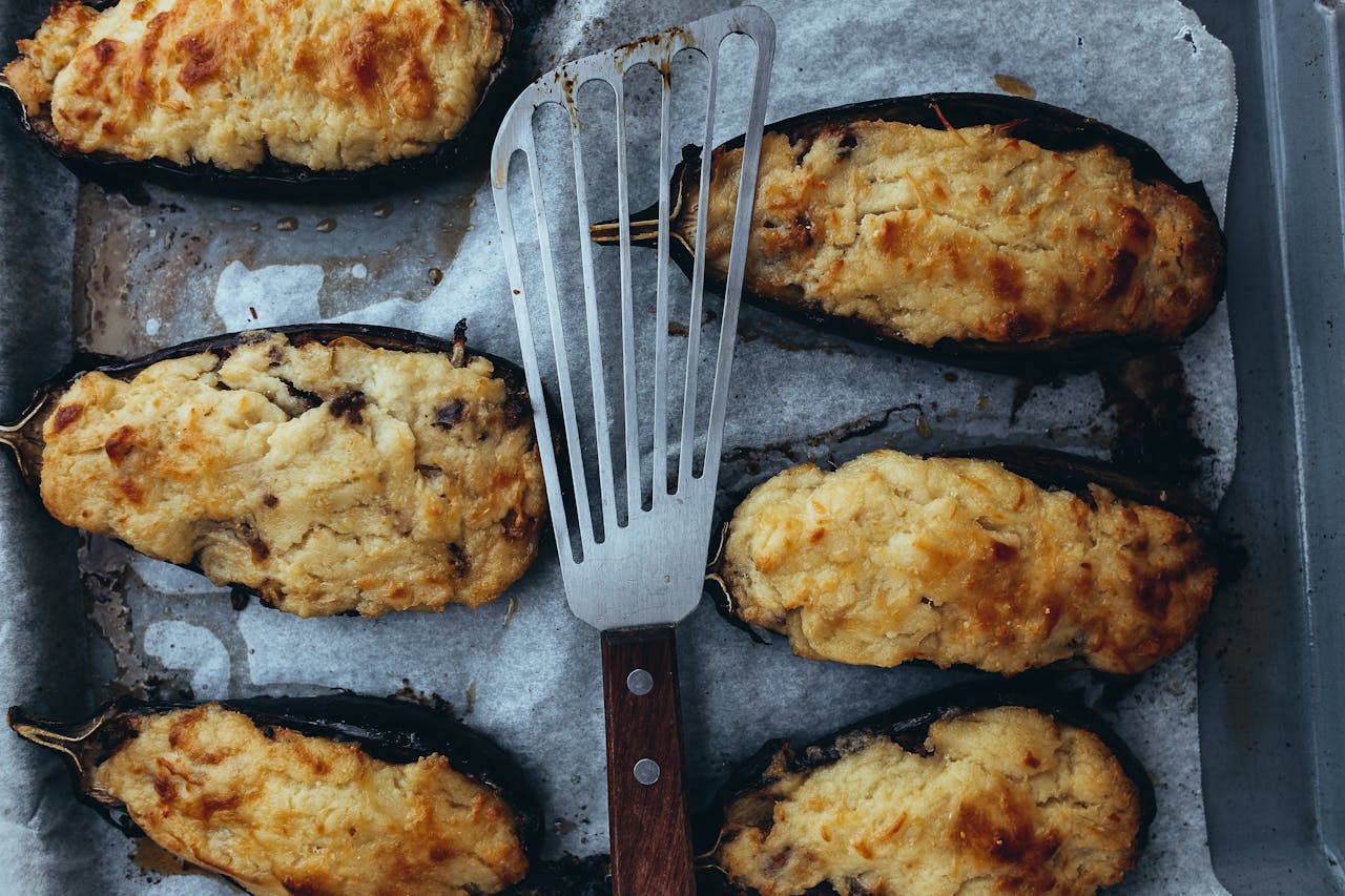 Baked stuffed eggplants with golden cheese topping on parchment-lined tray, metal spatula placed in center