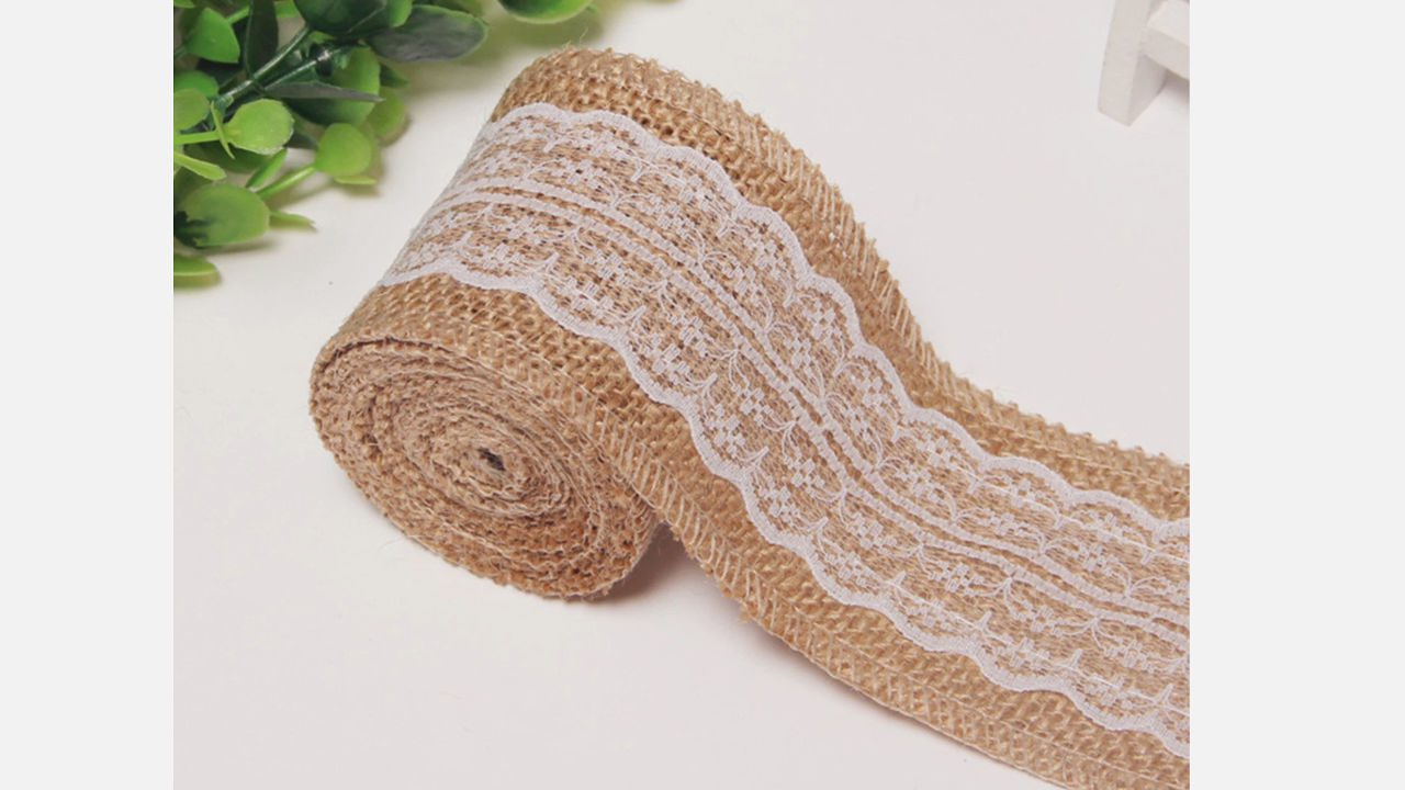 Roll of burlap ribbon with white lace trim, placed on a white surface near green artificial leaves