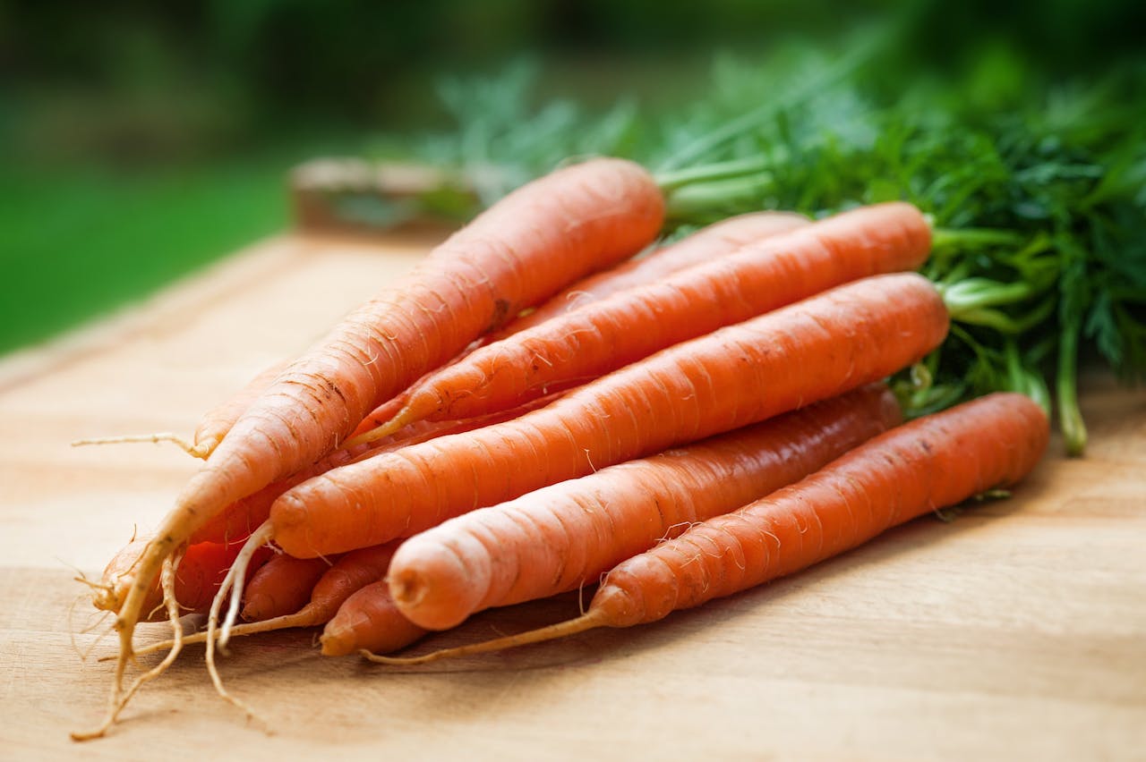 Freshly harvested carrots, placed on a wooden surface, with green leafy tops, outdoor natural lighting, close-up view, organic appearance, unpeeled and slightly dirty roots