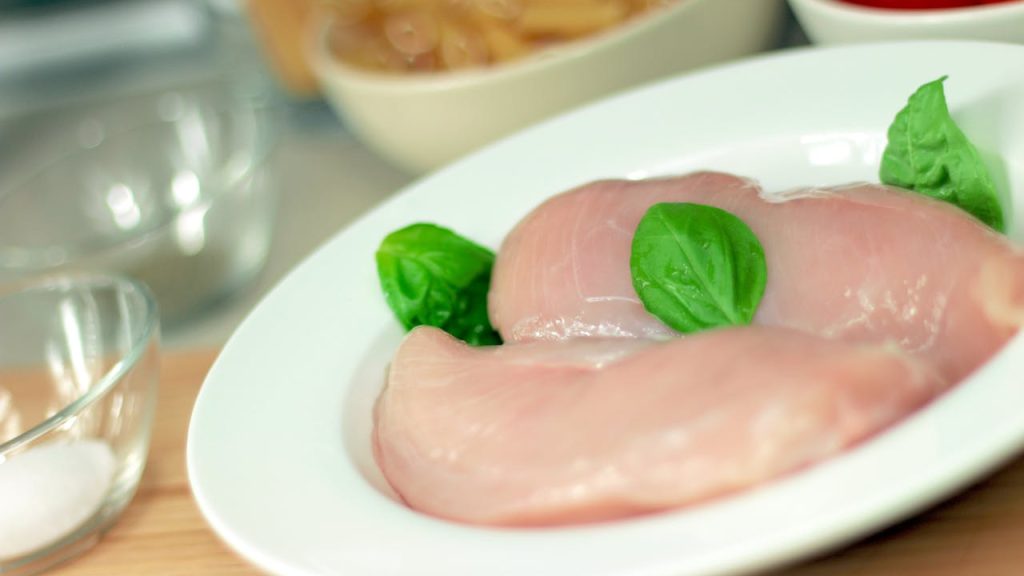 Raw chicken breasts on a white plate with basil leaves, pasta, tomatoes, and ingredients in background