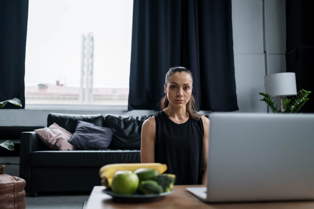 Woman sitting in front of a laptop in a living room with black curtains and a bowl of fruit on the table