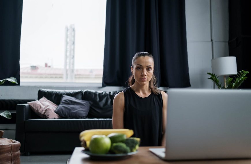 Woman sitting in front of a laptop in a living room with black curtains and a bowl of fruit on the table