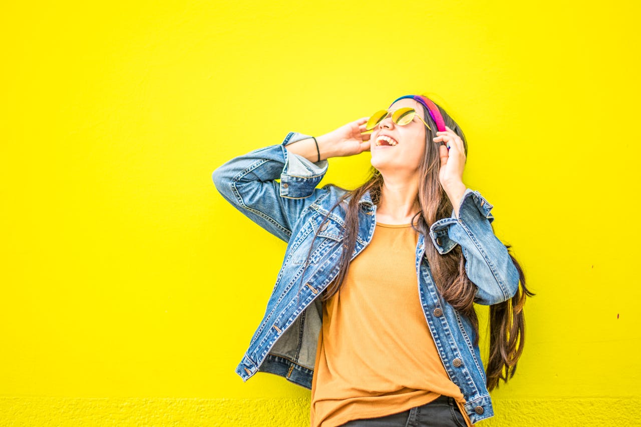 Smiling woman in a denim jacket and sunglasses standing against a bright yellow wall