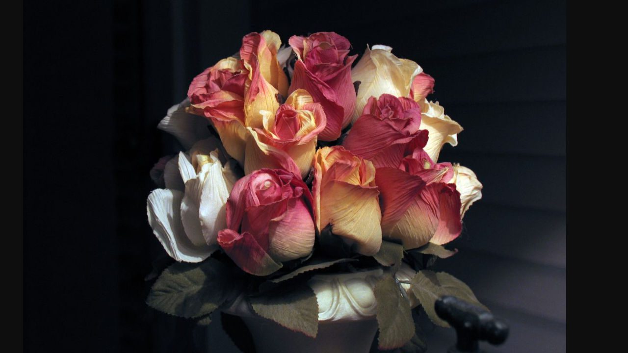 Artificial roses in shades of pink, peach, and cream arranged in a white vase under low dramatic lighting