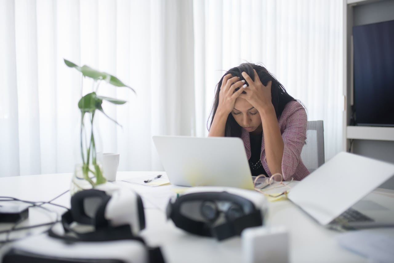 Frustrated woman holding head in hands, sitting at desk with laptop, VR headset, and notebooks, overwhelmed